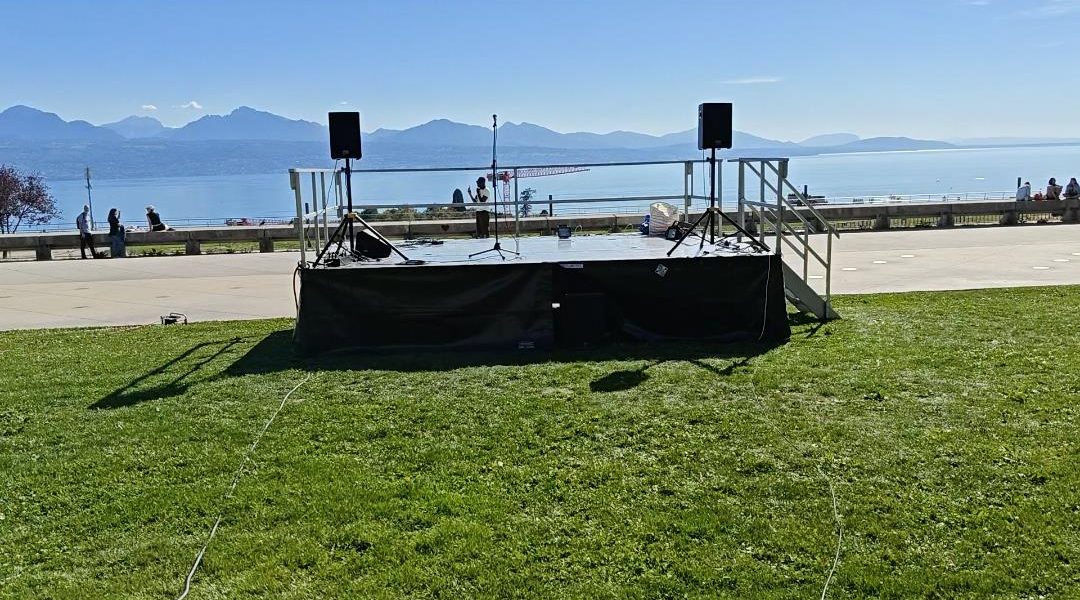 Scène extérieure avec deux enceintes sur pied, au bord du lac, montagnes en arrière-plan
